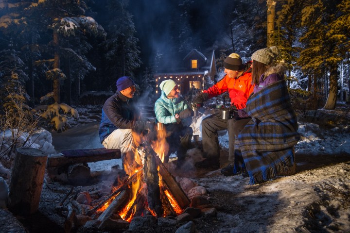 Group of four people sitting around a campfire in snowy forest at night near a lit cabin.