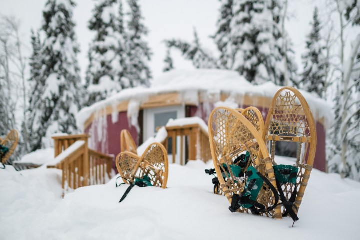 Snowshoes on snowy ground near a cabin with icicles, surrounded by snow-covered trees.